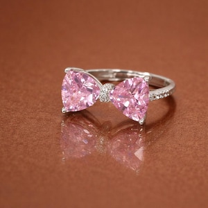 Könnte beinhalten: Ein silberner Ring mit einem Schleifendesign, das zwei große, herzförmige, rosa Edelsteine aufweist. Die Mitte der Schleife ist mit kleineren, klaren Steinen verziert. Der Ring liegt auf einer reflektierenden braunen Oberfläche.