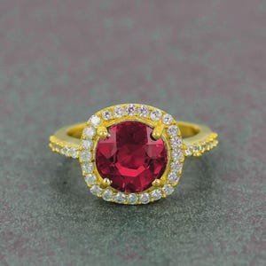 Puede incluir: Un anillo de oro con una gran piedra preciosa redonda de color rojo intenso, rodeada de piedras preciosas más pequeñas y transparentes. La banda del anillo también está adornada con pequeñas piedras preciosas transparentes. El anillo está sobre un fondo gris.