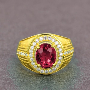 Può includere: Un anello d'oro con una grande gemma ovale rosso scuro al centro, circondata da un alone di piccole gemme trasparenti. La fascia dell'anello presenta un design strutturato con altre piccole gemme.