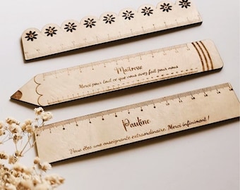 May include: Three wooden rulers with engraved messages. The top ruler has a scalloped edge with flower cutouts. The middle ruler is shaped like a pencil and says "Maitresse Merci pour tout ce que vous avez fait pour nous". The bottom ruler says "Pauline Vous êtes une enseignante extraordinaire. Merci infiniment!"