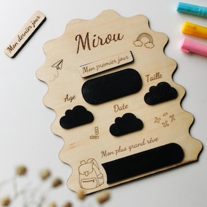 May include: A wooden milestone board with the name "Mirou" engraved at the top. It features spaces for "Age," "Date," and "Size," along with cloud and blackboard-shaped areas for writing. Includes the text "Mon premier jour" and "Mon plus grand rêve."