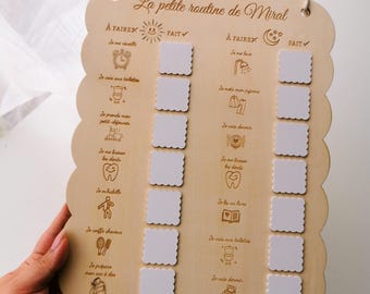 May include: A wooden daily routine chart with scalloped edges and the text "La petite routine de Miral." The chart has two columns with illustrated tasks and blank squares for check marks. A small hole is at the top for hanging.
