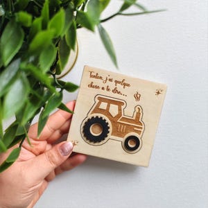 以下が含まれることがあります： トラクターのデザインと「Tonton, j'ai quelque chose à te dire...」の文字が入った木製の正方形。 トラクターは黒い車輪で、明るい色の木にエッチングされています。 手は緑の葉の近くで正方形を持っています。