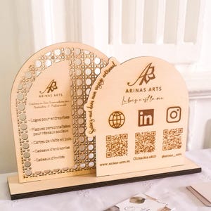 Peut inclure: Une enseigne en bois avec un design découpé au laser représentant une arche décorative et un logo pour Arinas Arts. L'enseigne comprend également le texte "Le bois c'est la vie" et des icônes de médias sociaux pour Instagram, LinkedIn et une adresse de site web.