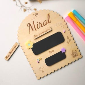 May include: A wooden chalkboard with the name "Miral" engraved at the top. It features two black rectangular chalkboards, decorative flowers, and the text "Mon premier jour" and "Date". Includes colorful markers.