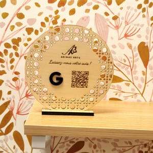 Peut inclure: Une pancarte ronde en bois avec un motif décoratif et un code QR. La pancarte porte le texte "ARINAS ARTS" et "Laissez-nous votre avis !". La pancarte porte également la lettre "G".