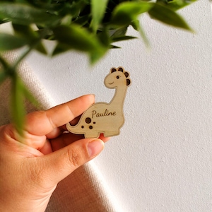 Puede incluir: Una etiqueta con forma de dinosaurio de madera con el nombre "Pauline" grabado. El dinosaurio tiene un cuello largo, una cara sonriente y manchas marrones. La etiqueta está hecha de madera de color claro.