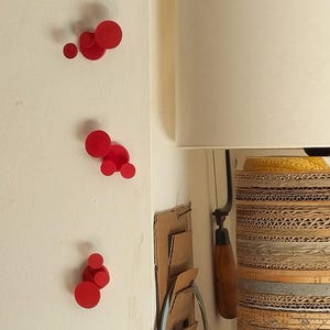 May include: A cylindrical lamp base constructed from stacked cardboard strips, topped with a cream-colored lampshade. Red circular wall decor is mounted on the adjacent white wall. A hand saw hangs nearby.