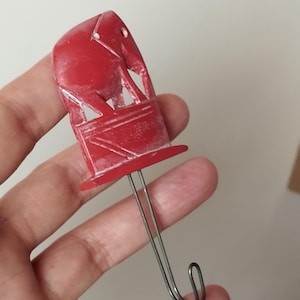 Op de afbeelding: Een rode, olifantvormige haak met een zilveren metalen draad. De olifant is gedetailleerd gesneden en heeft een klein gaatje aan de bovenkant. De haak wordt waarschijnlijk gebruikt om items op te hangen.