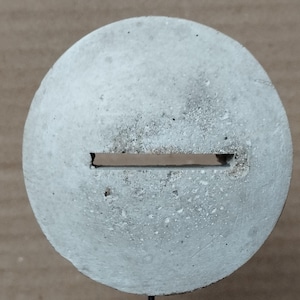 Può includere: Un disco rotondo in cemento grigio con una fessura orizzontale tagliata al centro. Il disco è appoggiato su un piccolo supporto nero.