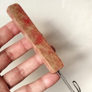 May include: A rectangular tool handle featuring a mottled tan and red marbled pattern. The handle is attached to a thin, silver-coloured metal hook extending from one end. The object has a smooth, flat surface with rounded edges and a uniform shape.