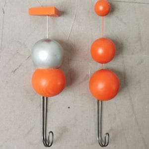 Puede incluir: Dos colgadores de pared decorativos. Uno presenta una parte superior rectangular naranja, una esfera plateada y una esfera naranja. El otro tiene tres esferas naranjas. Ambos tienen un gancho plateado en la parte inferior.