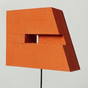 Puede incluir: Una escultura geométrica naranja con un recorte rectangular. La escultura tiene un diseño en capas con bordes angulados, montada sobre una fina varilla negra. La obra de arte está sobre un fondo blanco liso.