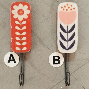 Peut inclure: Deux patères rectangulaires aux coins arrondis. La patère A présente un fond rouge avec un motif de fleur et de tige blanc. La patère B a un fond blanc avec un demi-cercle rose, des points orange et un motif de feuilles bleu foncé. Crochets en métal.