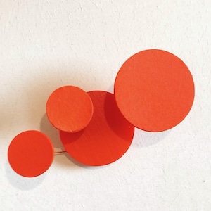 Può includere: Spilla arancione brillante composta da quattro cerchi di legno di diverse dimensioni, sovrapposti per creare un design moderno. La spilla è posta su uno sfondo bianco. Un accessorio di moda.