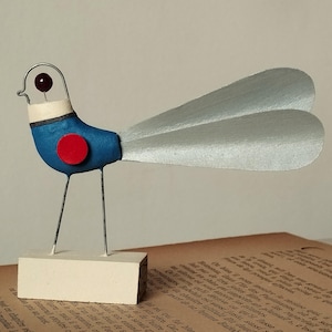 Puede incluir: Escultura de pájaro hecha a mano con cuerpo azul, alas plateadas y patas de alambre. Tiene un acento circular rojo y un cuello blanco. El ojo del pájaro es rojo oscuro. La escultura está montada sobre una pequeña base rectangular blanquecina.