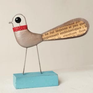 Könnte beinhalten: Eine silberne Vogelskulptur mit rotem und schwarzem Kopf, einem Schwanz aus Papier mit Text und auf einem blauen Holzsockel stehend.