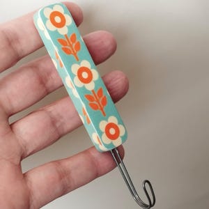 Peut inclure: Un crochet décoratif avec un manche rectangulaire turquoise. Le manche est orné d'un motif floral répété de fleurs blanches à cœur orange et de feuilles orange. Le crochet est en métal argenté.