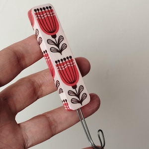 May include: A decorative hook with a rectangular, white body featuring a repeating pattern of red floral designs with black stems and leaves. The hook is made of metal.