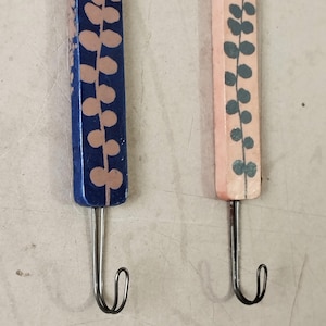 May include: Close-up of two wooden crochet hooks. One hook is painted navy blue with pink leaf accents, and the other is painted light pink with teal leaf accents. Each hook has a silver metal hook at the end.