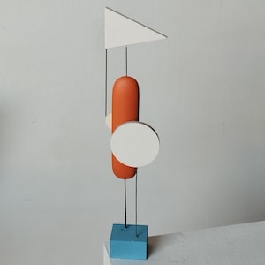 May include: Abstract wooden sculpture featuring a white triangle, orange oval, and white circle suspended by black wires. The sculpture rests on a blue square base. The design is minimalist and geometric.