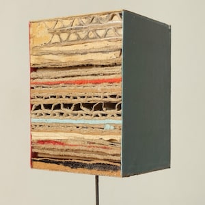 Puede incluir: Una escultura rectangular con una construcción de cartón en capas y un lado pintado de gris oscuro. Las capas expuestas revelan varios colores, incluyendo rojo y azul, dentro del cartón. La escultura está montada sobre una fina varilla metálica.