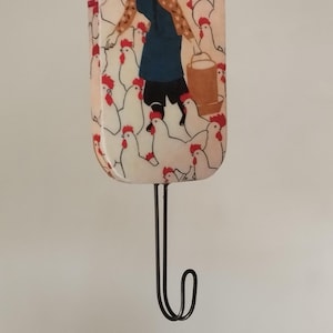 May include: A decorative wall hook featuring a whimsical illustration of a person with chickens. The rectangular hook has a black metal hook. The illustration depicts a person in a blue dress and a brown bucket surrounded by white chickens with red combs.