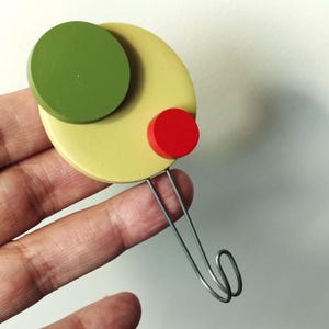 Op de afbeelding: Abstracte houten broche met een groen cirkel, een gele ovaal en een rode cirkel. De broche heeft een zilverkleurige draad haak aan de achterkant om aan kleding te bevestigen. Het ontwerp is modern en geometrisch.