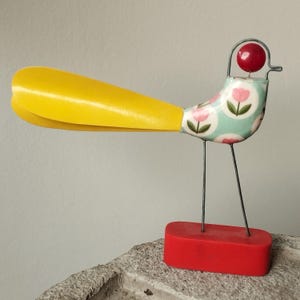 May include: A decorative bird figurine featuring a long yellow tail, a body patterned with pink tulips on a light blue background, a red spherical head, and thin metal wire legs standing on a solid rectangular red base.