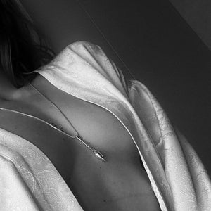 May include: A close-up of a person wearing a white robe with a silver pendant necklace. The pendant is a simple, elongated shape.