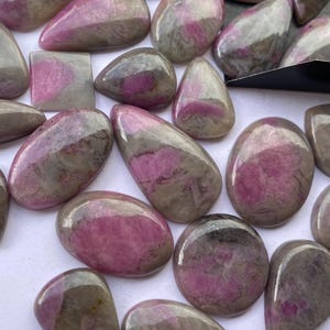 May include: A selection of polished gemstones in diverse forms, such as oval, teardrop, and rectangular. The stones display a blend of pink and grey tones, with a translucent appearance. The gemstones are arranged on a white surface.
