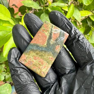 May include: A rectangular polished stone cabochon featuring a mottled pattern of earthy red, olive green, and dark charcoal grey mineral inclusions. The surface is smooth and reflective, displaying a natural, variegated texture across its flat face.