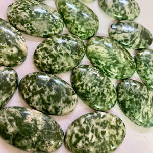 May include: A collection of polished green gemstones with a mottled pattern of darker green and white. The stones vary in shape, including oval and teardrop forms. The gemstones are arranged on a light-colored surface.