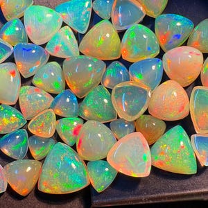 Può includere: Una collezione di pietre preziose di opale lucide in varie forme, tra cui triangoli e forme arrotondate. Gli opali mostrano un vibrante gioco di colori con sfumature di verde, blu, arancione e giallo, su una base bianco latte. Le pietre preziose sono disposte su una superficie scura.