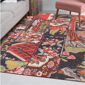 Op de afbeelding: Een kleurrijk patchwork tapijt met een bloemenmotief in tinten rood, groen, geel en zwart. Het tapijt ligt op een lichte houten vloer in een woonkamer.
