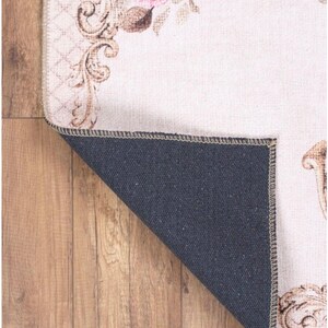 May include: A cream-colored rug with a floral design featuring a large red rose and intricate gold scrollwork. The rug has a black backing and is folded over to show the backing.