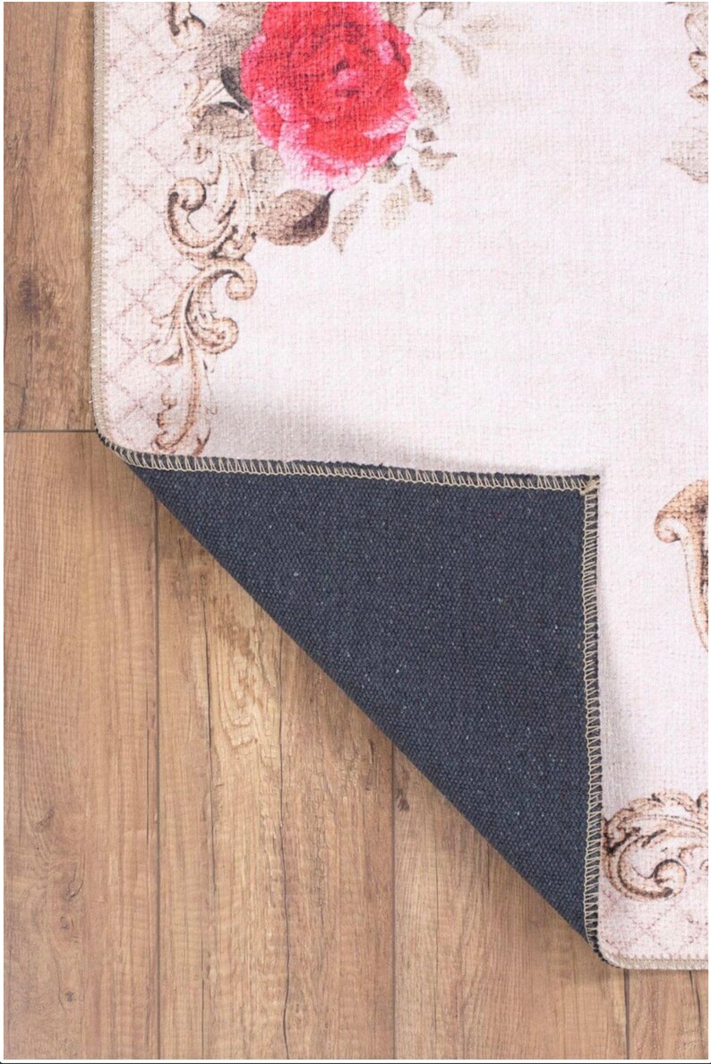May include: A cream-colored rug with a floral design featuring a large red rose and intricate gold scrollwork. The rug has a black backing and is folded over to show the backing.