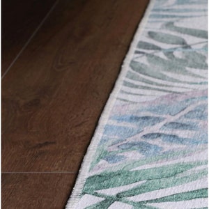 May include: A white rug with a green and blue floral pattern. The rug has a white border and is edged with a dark brown trim.