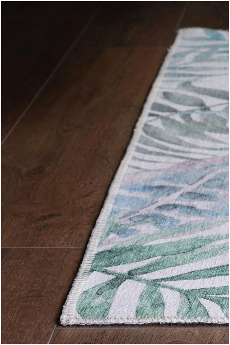 May include: A white rug with a green and blue floral pattern. The rug has a white border and is edged with a dark brown trim.