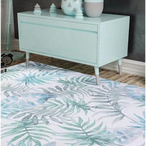 May include: A light blue dresser with a single drawer and wooden legs sits on a white rug with a green and blue watercolor leaf pattern. The dresser is decorated with a light blue vase, a gray vase, and three small gray and white ceramic trees.
