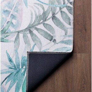 May include: A white rug with a watercolor design of green and blue leaves. The rug has a dark blue backing.