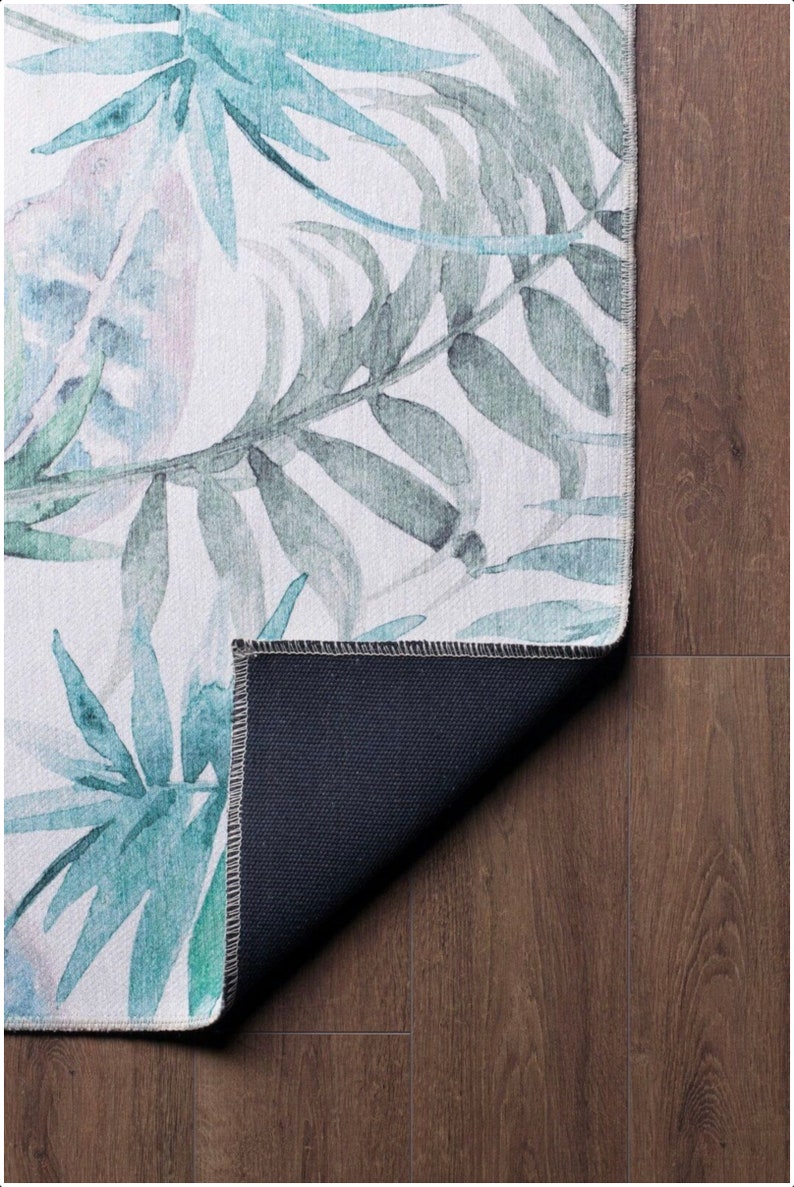 May include: A white rug with a watercolor design of green and blue leaves. The rug has a dark blue backing.