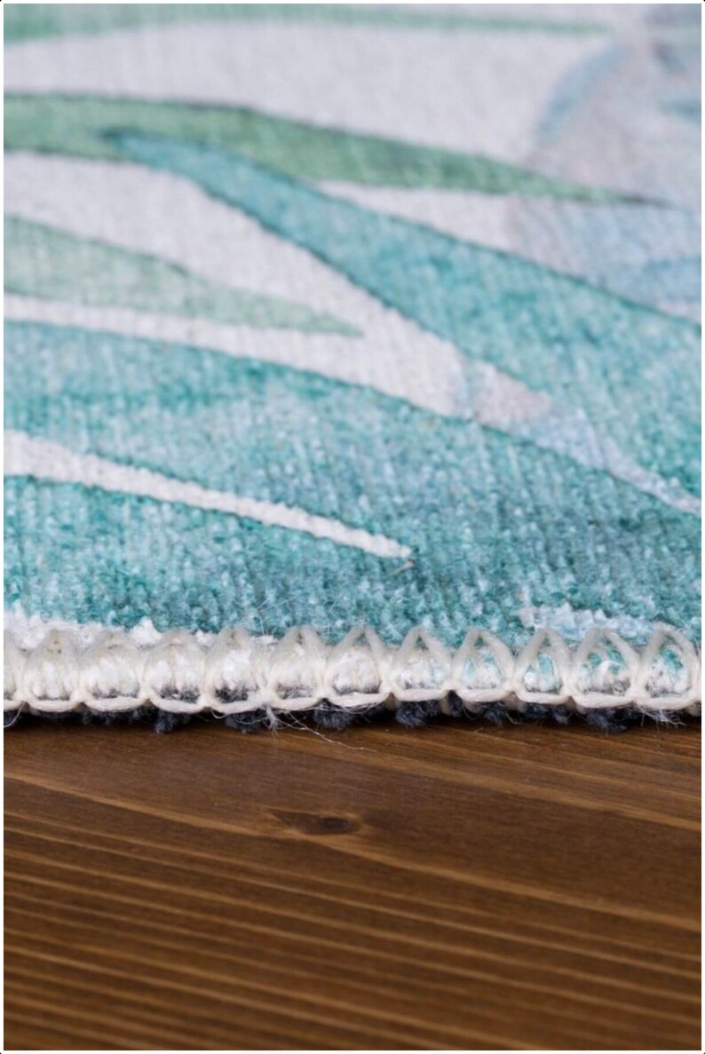 May include: Close-up of a teal and white patterned rug with a white fringe border. The rug is on a wooden surface.