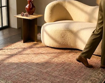May include: A rectangular area rug featuring a faded, distressed pattern in shades of muted red, pink, and sage green with a wide, intricate border. The rug is positioned on a dark wood floor, partially underneath a cream-colored, curved sofa.