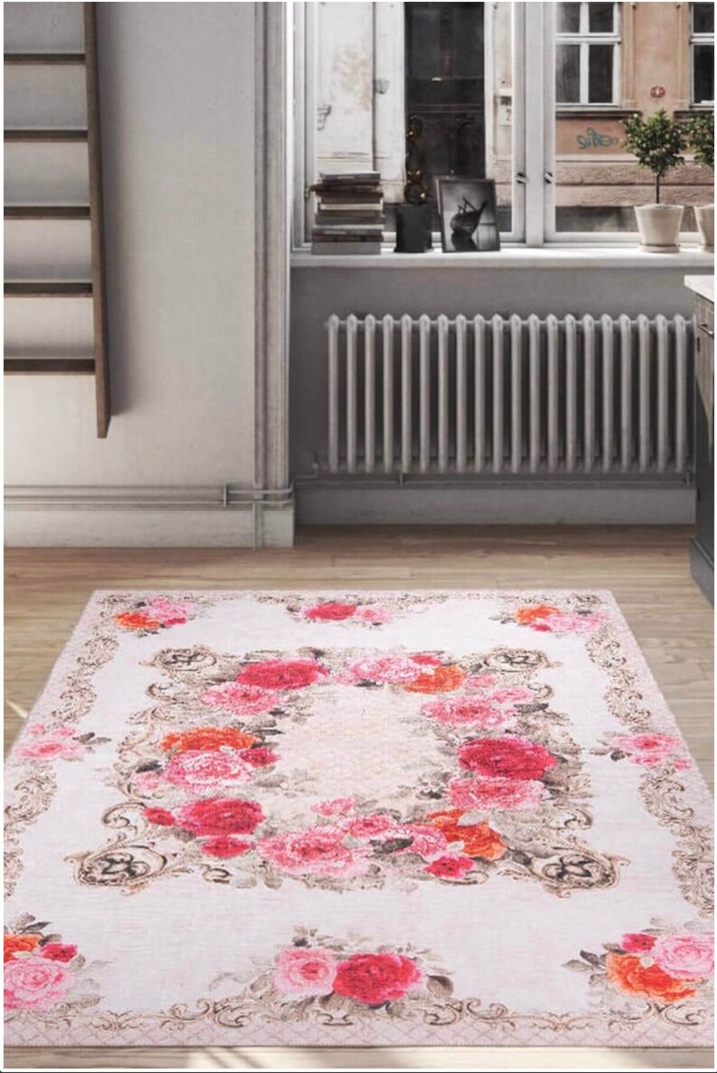 May include: A white rug with a floral pattern featuring pink, red, and orange roses. The rug has a decorative border with a gold-colored design.