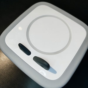 Könnte beinhalten: Ein quadratisches, weißes und graues Gerät mit einer kreisförmigen Vertiefung und zwei schwarzen, oval geformten Knöpfen. Das Gerät hat abgerundete Ecken und ein glattes Design. Der Hintergrund ist dunkel.