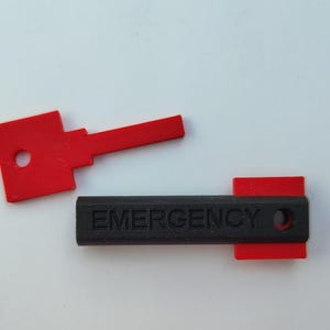 Op de afbeelding: Een zwarte kunststof sleutel met het woord "EMERGENCY" erop gegraveerd. De sleutel heeft aan beide uiteinden een rode kunststof dop.