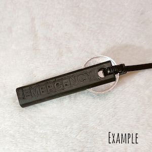 Peut inclure: Un porte-clés en plastique noir avec le mot "EMERGENCY" en relief. Le porte-clés est attaché à un disque en métal argenté et à un cordon noir.
