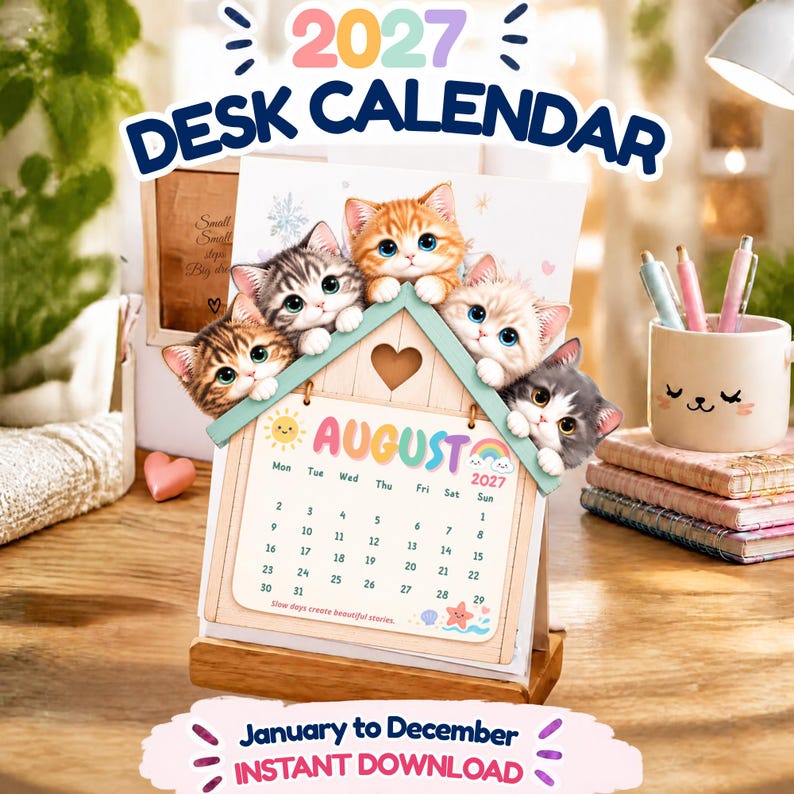 Puede incluir: Un calendario de escritorio 2027 con un marco en forma de casa y cuatro gatitos ilustrados asom&aacute;ndose por el tejado. La p&aacute;gina del calendario muestra el mes de agosto con un sol, un arco&iacute;ris y una estrella de mar. Texto: 2027 DESK CALENDAR.