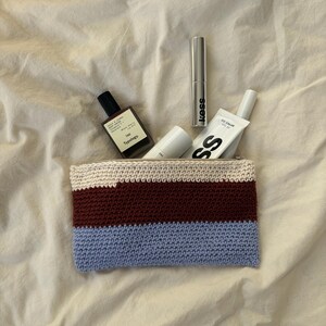 Könnte beinhalten: Eine gestreifte Kosmetiktasche aus Häkelarbeit in Creme, Burgunderrot und Hellblau, die Schönheitsprodukte enthält. Die Tasche enthält eine Parfümflasche, Mascara und Hautpflegetuben. Die Artikel sind auf einem cremefarbenen Stoff angeordnet.